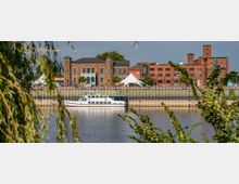 Historisches Backsteingebäude und moderner Pavillon direkt am Ufer eines Flusses, umgeben von Grünflächen. Ein weißes Ausflugsboot liegt am Kai, eingerahmt von Baumästen im Vordergrund.