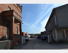 Industriegelände mit zwei gegenüberliegenden Gebäuden, eines aus rotem Backstein und das andere in grauem Putz. Auf dem betonierten Hof sind geparkte Fahrzeuge und einige Baustellenmaterialien sichtbar, im Hintergrund weitere Industriehallen und blauer Himmel.