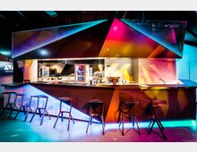 Ein moderner Barbereich mit futuristischem Design, beleuchtet durch bunte, geometrische Lichtprojektionen. Die Bar verfügt über hohe, minimalistische Hocker, eine Theke mit Getränkespendern und Kaffeemaschine sowie ein Regal mit Red-Bull-Dosen im Hintergrund.