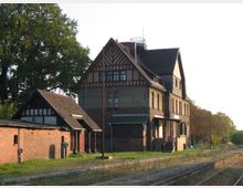 Ehemaliges Bahnhofsgebäude aus Backstein mit Fachwerkelementen, umgeben von niedrigen Nebengebäuden und einer stillgelegten Bahnstrecke; im Hintergrund stehen große Bäume und eine Baumreihe entlang des Gleises.
