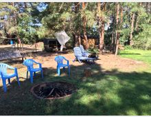 Offene Wiese mit Kiefern und Gartenmöbeln, darunter blaue Plastikstühle und Liegen, die um eine Feuerstelle aus Steinen gruppiert sind. Im Hintergrund befinden sich ein Schuppen, gestapeltes Holz und eine Schaukel unter den Bäumen.