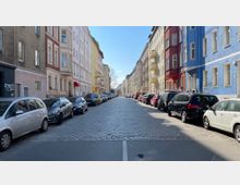 Straßenansicht mit mehrstöckigen, bunt gestrichenen Wohnhäusern auf beiden Seiten der gepflasterten Straße. Autos parken in Reihen entlang der Bordsteine unter einem klaren, blauen Himmel.
