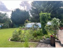Garten mit gepflegtem Rasen, Blumenbeeten und großen immergrünen Bäumen im Hintergrund. Im Zentrum ein rankender Pflanzenbogen, rechts eine Terrasse mit Blumen in Töpfen und links ein Wäschegestell sowie eine kleine grüne Gartenhütte.