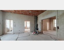 Innenraum einer Baustelle mit rohe verputzten Wänden, offenliegenden Kabeln und einem Betonboden. Fenster lassen Tageslicht herein, und ein alter Kamin sowie Baumaterialien wie eine Schubkarre und Säcke sind sichtbar.