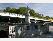 Steinbrücke mit einer Statue auf einem Sockel, die eine stehende Figur mit erhobenem Arm zeigt. Im Hintergrund eine erhöhte Bahnlinie mit einer gelben U-Bahn, dahinter Bäume und modernes Gebäude.