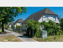 Eine ruhige Wohnstraße mit Einfamilienhäusern, die weiße Fassaden und braune Schrägdächer haben, umgeben von Gärten mit viel Grün. Ein gepflasterter Gehweg verläuft entlang eines Zauns, hinter dem die Häuser stehen, und ein Straßenschild mit der Aufschrift „Freiligrathstraße“ ist sichtbar.