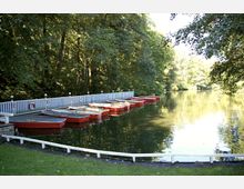 Bootsanlegestelle mit mehreren roten Holzbooten, die an einem Steg festgemacht sind, umgeben von dichtem, grünem Wald und ruhigem Wasser. Im Vordergrund befindet sich eine grasbewachsene Fläche mit einem weißen Geländer.