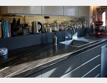 Moderne Küche mit dunkler Arbeitsplatte aus Marmoroptik, einer eingebauten Spüle und Wasserhahn, darüber ein Regal mit Einmachgläsern an einer Wand aus freigelegtem Backstein. Auf der Arbeitsfläche stehen Küchengeräte, Bücher und Zubehör wie ein Wasserkocher und ein Messerblock.