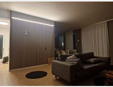 Modernes Wohnzimmer mit Holzelementen: Ein Raum mit heller Holzvertäfelung an der Wand, einer grauen Couch mit Kissen und dunklem Teppichboden. Im Hintergrund große Fenster mit weißen Vorhängen und ein Esstisch mit Stühlen.