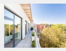 Moderne Dachterrasse mit Glasgeländer, großen bodentiefen Fenstern und hellen Fliesen, umgeben von Bäumen und mit Blick auf rote Ziegeldächer benachbarter Gebäude unter klarem Himmel.
