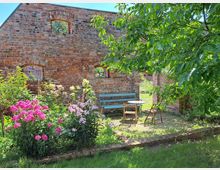 Backsteinmauer einer Ruine mit mehreren offenen Fenstern und einem begrünten Garten im Vordergrund; bunte Blumenbeete, eine Holzbank und ein kleiner Tisch mit Stühlen stehen im Schatten eines Baums.