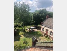 Blick auf einen historischen Hof mit Ziegelgebäuden, Kopfsteinpflaster und einer Wiese, umgeben von hohen Bäumen. Im Hof steht eine überdachte Sitzgruppe, im Hintergrund Wäschestücke an einer Leine.