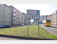 Straßenkreuzung in einer Stadt mit einem Verkehrszeichen, das verschiedene Abbiegeoptionen Richtung "Zentrum" und "Kurfürstendamm" anzeigt, umgeben von mehrstöckigen Wohngebäuden. Im Vordergrund eine begrünte Verkehrsinsel und im Hintergrund ein gelber Doppeldeckerbus auf der Straße.