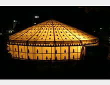 Ein rundes, kuppelförmiges Zelt mit leuchtend gelber Beleuchtung, geschmückt mit einem Muster aus dunklen Verzierungen auf der Oberfläche, steht in einer nächtlichen Umgebung. Im Hintergrund sind schwach beleuchtete Gebäude und Straßenlaternen zu erkennen.