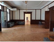 Ein Raum mit Parkettboden und Wänden, die teilweise mit dunklem Holz vertäfelt und weiß gestrichen sind. Im Vordergrund befinden sich Sitzmöbel, im Hintergrund führt der Raum zu einem Bereich mit Holztischen und Stühlen.