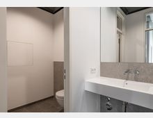 Modernes Badezimmer mit weißem Waschbecken auf grauen Fliesen, integriertem Wasserhahn und einem großen Spiegel. Im Hintergrund ist eine Toilettenschüssel vor einer halbhohen gefliesten Wand zu sehen.