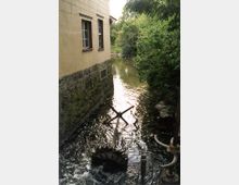 Ein kleines Flussbett mit klarem Wasser, flankiert von einer Steinmauer eines Gebäudes mit Fenstern links und dichter grüner Vegetation rechts. Im Wasser befindet sich ein altes Wasserrad, das teilweise sichtbar ist.