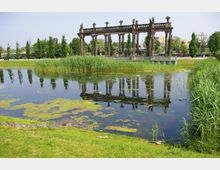 Teichlandschaft mit grasbewachsenen Ufern und hohen Schilfpflanzen, im Hintergrund eine historische Steinsäulenkonstruktion mit Skulpturen, deren Spiegelbild im ruhigen Wasser sichtbar ist.