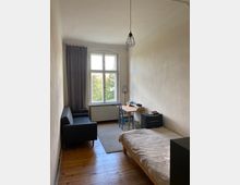 Schmaler Raum mit Holzboden, einem Einzelbett rechts, einem kleinen Schreibtisch vor einem Fenster mit grauem Vorhang sowie einem schwarzen Sofa links. An der Decke hängt eine einfache Lampe mit Metallgitter.