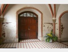 Innenansicht eines historischen Gebäudes mit einem hölzernen Doppeltor in einem gemauerten Rundbogen. Die Wände sind teilweise verputzt und weisen Abblätterungen auf, der Boden ist mit rot-weiß gemusterten Fliesen belegt, und ein Blumenarrangement in einem weißen Gefäß steht rechts vor der Wand.