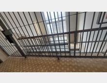 Treppenhaus mit Metallgitter-Abtrennung im Vordergrund, dahinter abwärts führende Treppen mit Stein- und Metallgeländern sowie ein großes Fenster mit strukturiertem Glas, das Tageslicht hineinlässt. Wände und Boden mit schlichten Designs, darunter ein gemustertes Fliesenmuster.