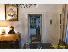 Esszimmer mit Holztisch und Stühlen, einer Kommode mit dekorativer Tischlampe und antiker Uhr. Eine Tür im Hintergrund führt in eine helle Küche mit weißen Schränken und einigen Küchenutensilien.