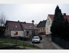 Straßenszene in einer Wohnsiedlung mit Einfamilienhäusern, charakterisiert durch rote Ziegeldächer und gepflasterte Wege. Im Vordergrund steht ein geparktes Auto, und im Hintergrund sind ein Baum sowie weitere Häuser sichtbar.
