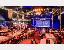 Festlich dekorierter Veranstaltungsraum mit einer Bühne im Vordergrund, umgeben von glitzernden Vorhängen und Lichterketten. Der Raum ist mit Tischen und Stühlen ausgestattet, die für ein Essen gedeckt sind, unter mehreren eleganten Kronleuchtern.