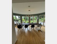 Großes Esszimmer mit einem langen Holztisch und verschiedenen Stühlen auf einem hellen Holzboden. Der Raum ist von bodentiefen Fenstern umgeben, die einen Blick auf einen Garten mit Bäumen und Rasen bieten; eine moderne Hängeleuchte hängt über dem Tisch.