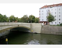 Straßenbrücke aus Beton mit dekorativen Elementen, die über einen Fluss führt; im Hintergrund ein mehrstöckiges Wohngebäude sowie grüne Bäume und Baustellenfahrzeuge. Der Fluss hat grünlich schimmerndes Wasser, und am Ufer ist eine gemauerte Böschung sichtbar.