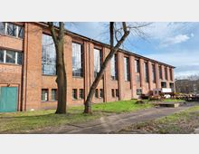 Backstein-Gebäude mit großen vertikalen Fenstern, flankiert von Bäumen und umgeben von einer Grünfläche. Im Vordergrund befinden sich Gehwege, während rechts im Bild Teile von industriellen Geräten zu sehen sind.