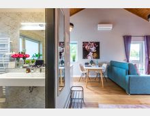 Wohnraum mit angrenzendem Badezimmer in einem modernen, hellen Interieur. Der Raum enthält ein Sofa, einen Esstisch mit Stühlen, ein großes Wandbild und Fenster mit lila Vorhängen; das Badezimmer ist mit Marmoroptik-Wänden und Waschbecken ausgestattet.