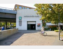 Eingang zum S-Bahnhof "Innsbrucker Platz" mit weißer Fassade, Schriftzug und S-Bahn-Logo, daneben ein Marktstand unter einem gestreiften Markisendach. Im Hintergrund fährt eine gelbe S-Bahn über eine erhöhte Brücke, im Vordergrund ein gepflasterter Platz mit einem Mann und einem Fahrrad.