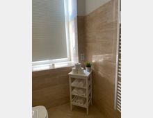 Ein helles Badezimmer mit beigen Fliesen an den Wänden und auf dem Boden. Ein kleines weißes Regal steht in der Ecke, darauf liegen gerollte Handtücher, Toilettenpapierrollen und eine kleine Pflanze im Topf. Rechts im Bild ist ein weißer Handtuchheizkörper, und links ist ein Fenster mit geschlossener Jalousie.