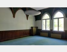 Leerer Raum mit neogotischen Spitzbogenfenstern, verziertem Holzwandpaneel und gemusterten Deckenbögen. Der Boden ist mit einem türkisfarbenen Teppich ausgelegt, und außen sind Bäume durch die Fenster sichtbar.