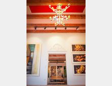 Innenraum mit moderner Einrichtung, einer auffälligen, bunten Deckenleuchte, Holzbalken an der Decke und Wandverzierungen. Im Vordergrund eine Türöffnung, die zu einem Raum mit Holztisch, Stühlen und einer Bücherwand führt; an den Wänden hängen großformatige Kunstwerke.