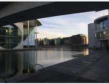 Architekturkomplex am Spreeufer in Berlin mit modernen Gebäuden aus Glas und Beton, darunter das Marie-Elisabeth-Lüders-Haus mit einem markanten runden Fenster. Im Vordergrund verläuft die Spree, ergänzt durch breite, gepflasterte Fußwege.