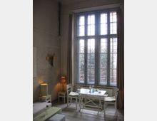 Helles Wohnzimmer mit hohen, vergitterten Fensterfronten und cremefarbenen Vorhängen. Im Raum stehen ein weißer Tisch mit Stühlen, eine Sitzbank, zwei Lampen und Dekorationselemente auf hellen Holzböden vor einer unverputzten Backsteinwand.