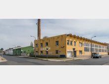 Ecke einer städtischen Straße mit einem zweistöckigen gelben Industriegebäude mit gezacktem Dach und großen Fenstern, daneben ein Schornstein aus Backstein. Im Hintergrund ein grünes Lagergebäude und Wohnhäuser.