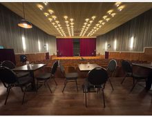 Ein Theatersaal mit Reihen von Holzstühlen und kleinen Tischen mit Stühlen im Vordergrund. An der Stirnseite befindet sich eine Bühne mit einem geschlossenen roten Vorhang, umgeben von hölzernen Wandverkleidungen und Wandlampen; die Decke ist mit dekorativen Leuchten ausgestattet.