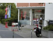 Straßenszene mit einem Café mit großen Glasfenstern und einer Leuchtschrift „Café“ über dem Eingang. Davor stehen ein Fahrrad, eine Motocross-Maschine und einige rot-weiße Stehtische; daneben befinden sich bunte Fahnen und ein Straßenschild mit der Aufschrift „Altonaer Straße“.