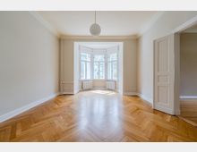 Heller, leerer Altbau-Wohnraum mit Fischgrätparkett, hohen Decken mit Stuck und einer zentralen Pendelleuchte. Am Ende des Raums befindet sich ein großer Erker mit mehreren Fenstern und Heizkörpern darunter; rechts ist eine offene Tür zu einem angrenzenden Zimmer zu sehen.