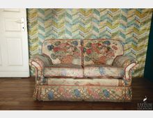 Ein gepolstertes Zweisitzersofa mit floralen Mustern in Pastellfarben steht vor einer Wand mit einem bunten, grafischen Zickzackmuster. Der Boden besteht aus hellem Holz, und links ist eine weiße Tür zu sehen.