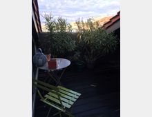 Kleine Dachterrasse mit Holzplankenboden, mehreren Pflanzen in Töpfen, einem klappbaren grünen Stuhl und einem kleinen runden Tisch mit Dekoelementen. Im Hintergrund sind Dächer und ein teilweise wolkiger Himmel sichtbar.