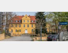 Gelbes, zweistöckiges Gebäude mit einem roten Satteldach und Dachgauben, umgeben von Bäumen und einer gepflasterten Zugangsstraße. Im Vordergrund befinden sich geparkte Autos und ein Straßenschild mit der Aufschrift „Weberweg“.