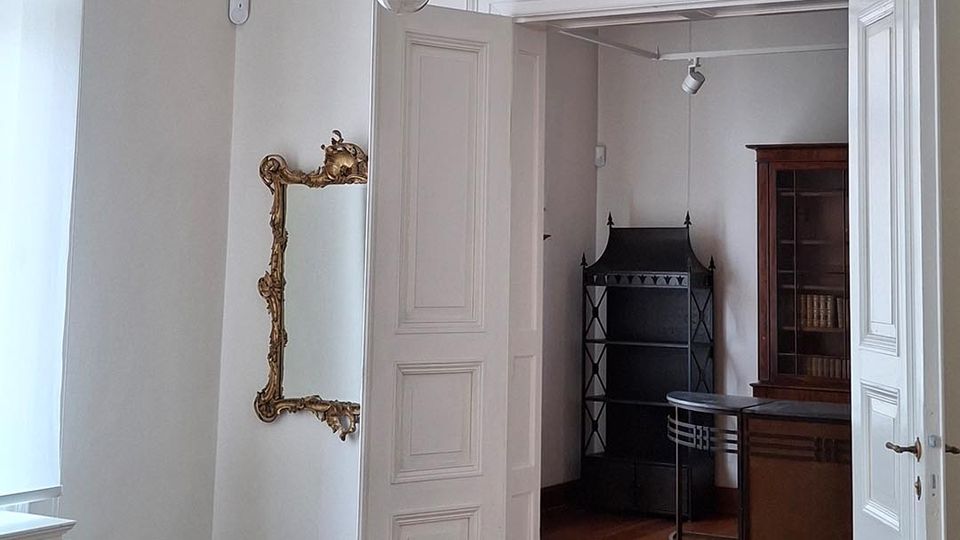 Heller Altbau-Innenraum mit hohen Decken, Stuckfries und großem Kristalllüster über Holzdielenboden. Offene doppelflügelige Tür führt in ein angrenzendes Zimmer mit Vitrine und kleinem Tisch; an der Wand hängt ein goldgerahmter Spiegel.