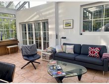 Wohnzimmer mit großen Fensterfronten und Oberlichtern, die viel Tageslicht hereinlassen. Der Raum enthält eine dunkelblaue Couch mit bunten Kissen, einen modernen Sessel, einen Glastisch, ein Klavier und Fliesenboden in terrakottafarbenem Muster.