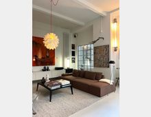 Modern living room with high ceilings and exposed beams, featuring a brown sectional sofa on a large rug and a black coffee table stacked with books. White walls display large abstract artwork, a sculptural pendant light hangs overhead, and a gridded glass door and tall radiator sit along the back wall.