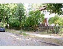 Gepflasterte Straße mit einem Einfamilienhaus im Hintergrund, das von einem Holzzaun und dichtem grünen Laub umgeben ist. Vor dem Haus führen ein Fußweg und eine kleine Eingangstreppe zur Haustür.