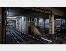 Ein U-Bahn-Tunnel mit mehreren Gleisen, einem kleinen gemauerten Betriebsraum mit Graffiti und einer Metalltreppe. Die Umgebung wirkt industriell, mit Kabeln und Beleuchtungslampen an den Decken und Wänden.